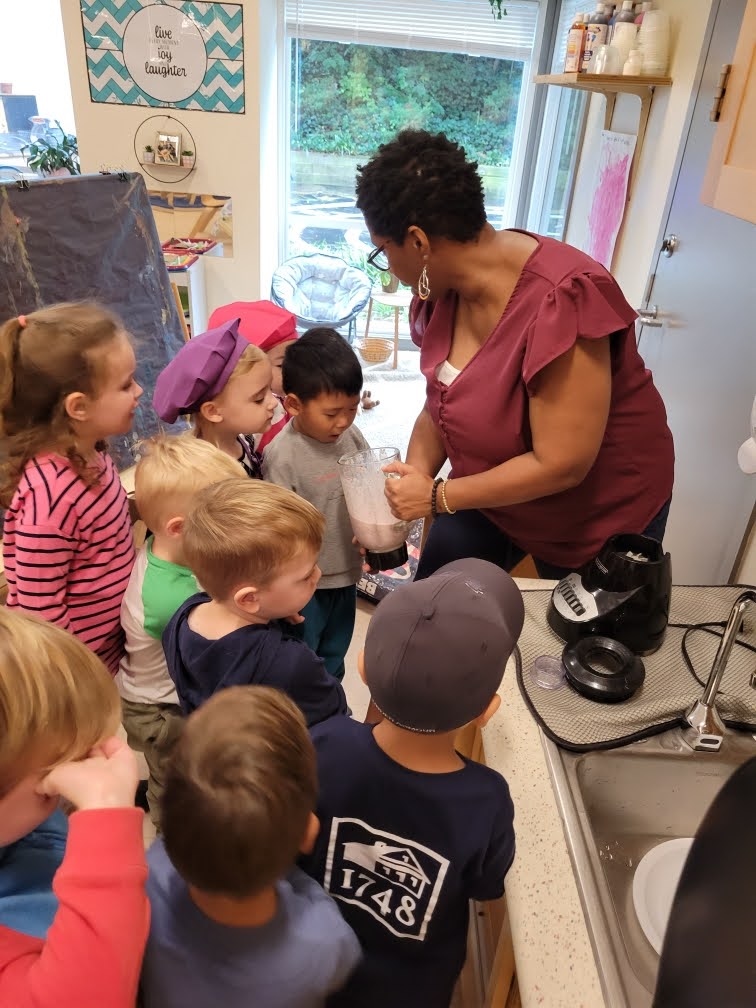 Pilot Light educator Lisa M. shows her Pre-K students how to make a delicious smoothie. A group of about seven students gathers around Lisa as she shows them a blender with light pink liquid inside. 