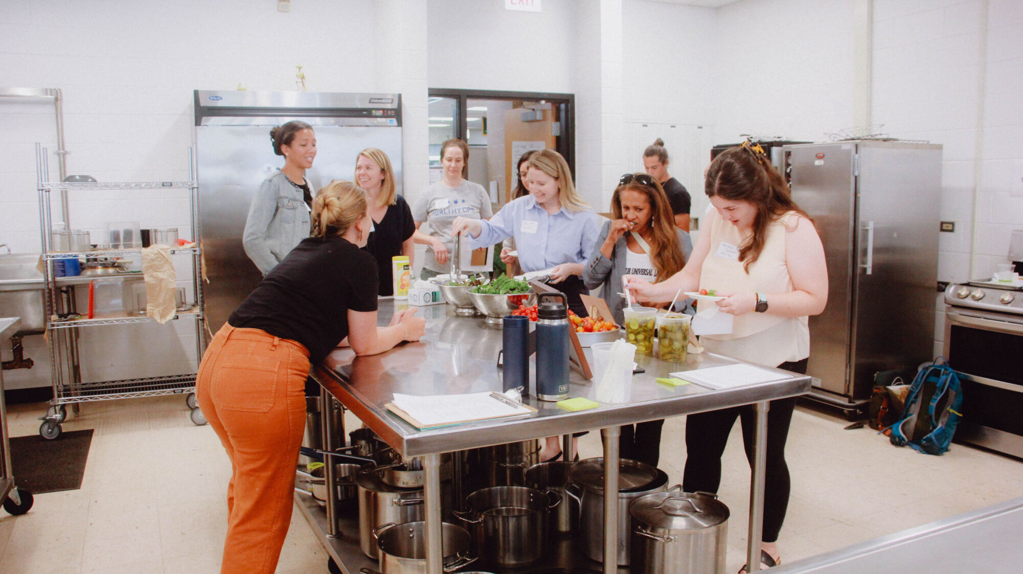 A group of seven adults in a classroom, taste testing tomatoes, lettuce, and pickles as part of an agriculture education lesson demonstration.