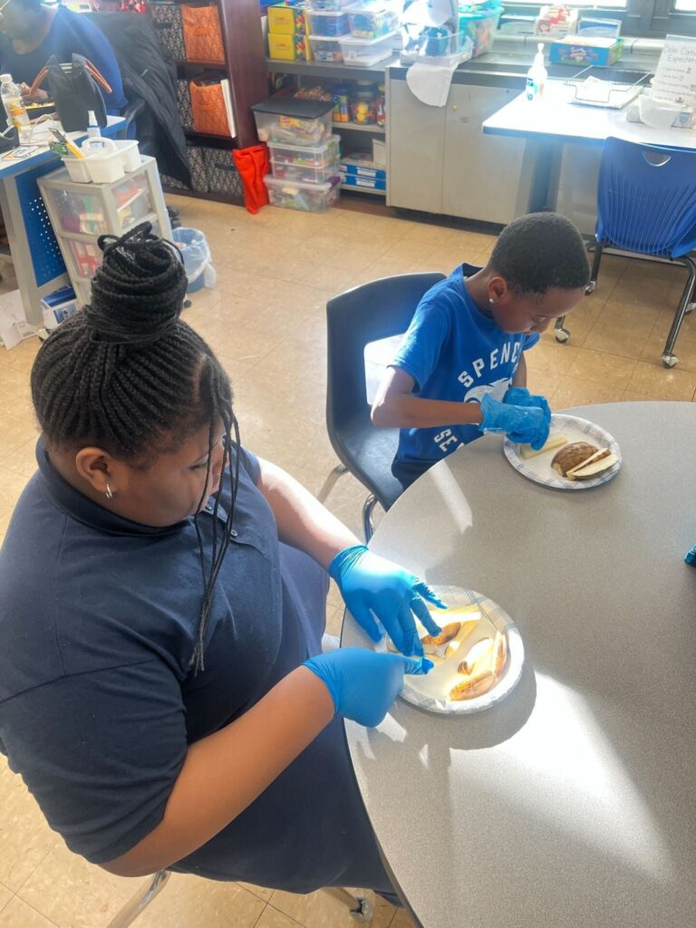 Two elementary students wearing sanitary gloves carefully slice potatoes to make homemade french fries in their classroom air fryer.