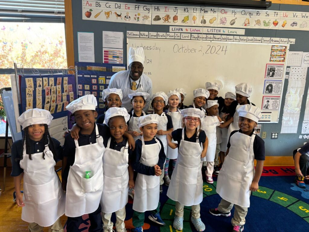 A teacher stands with a group of elementary aged students all wearing chefs hats and looking into the camera.