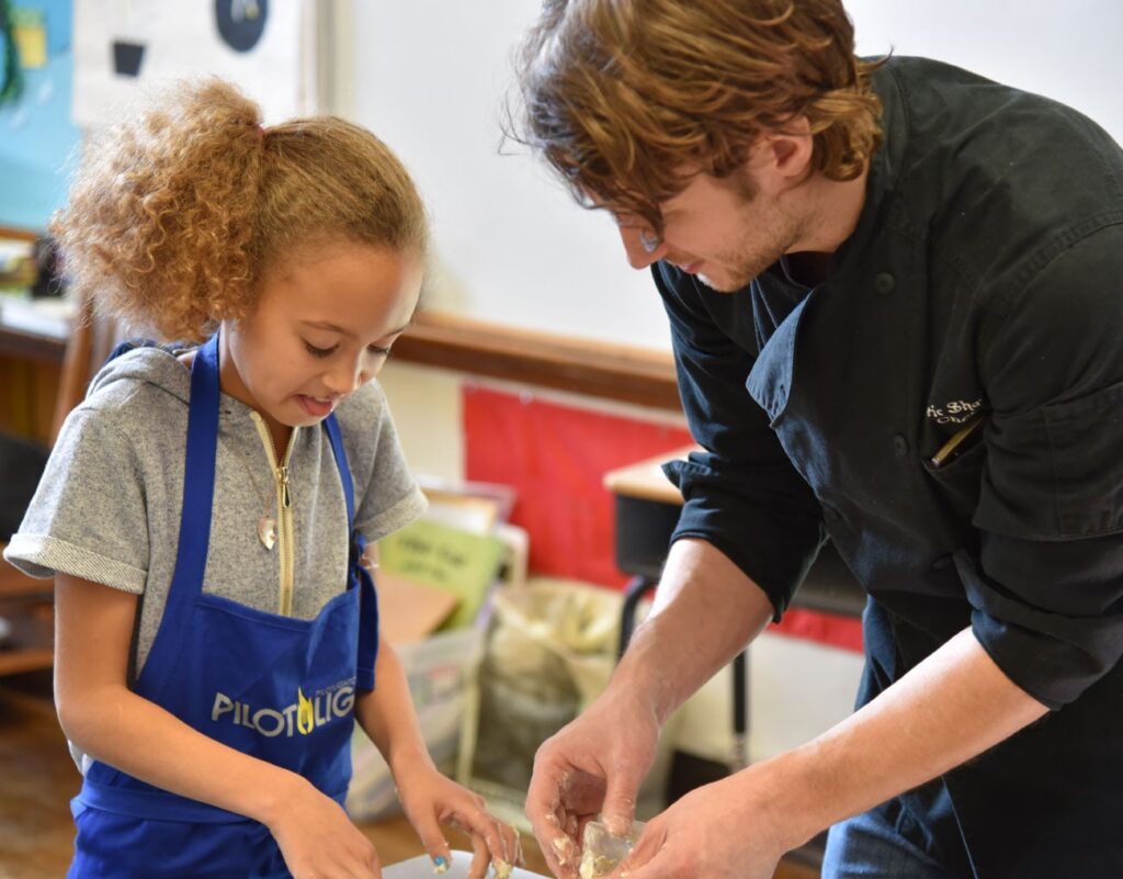 Young girl in a blue apron learns hands-on cooking from a chef in a classroom kitchen, creating a warm, focused moment.