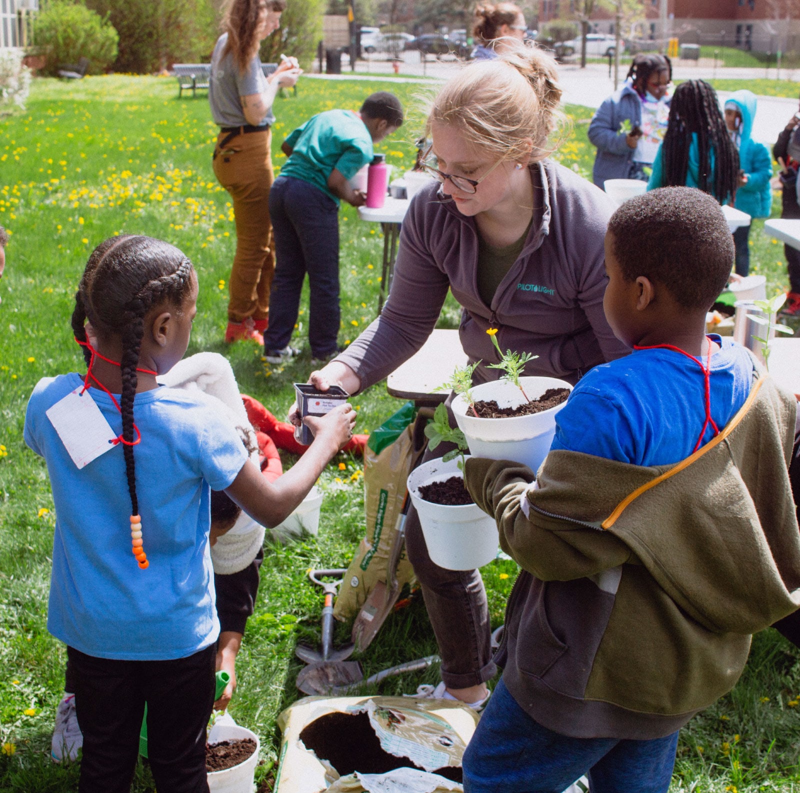 Children and an adult volunteer plant seedlings in pots during a sunny community gardening event, creating a cheerful, collaborative scene.