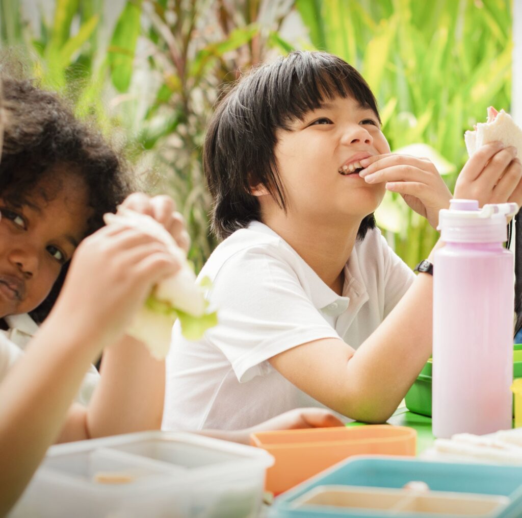 Two children enjoy lunch outdoors: one child bites a sandwich while another eats nearby, with a pink water bottle on the table.