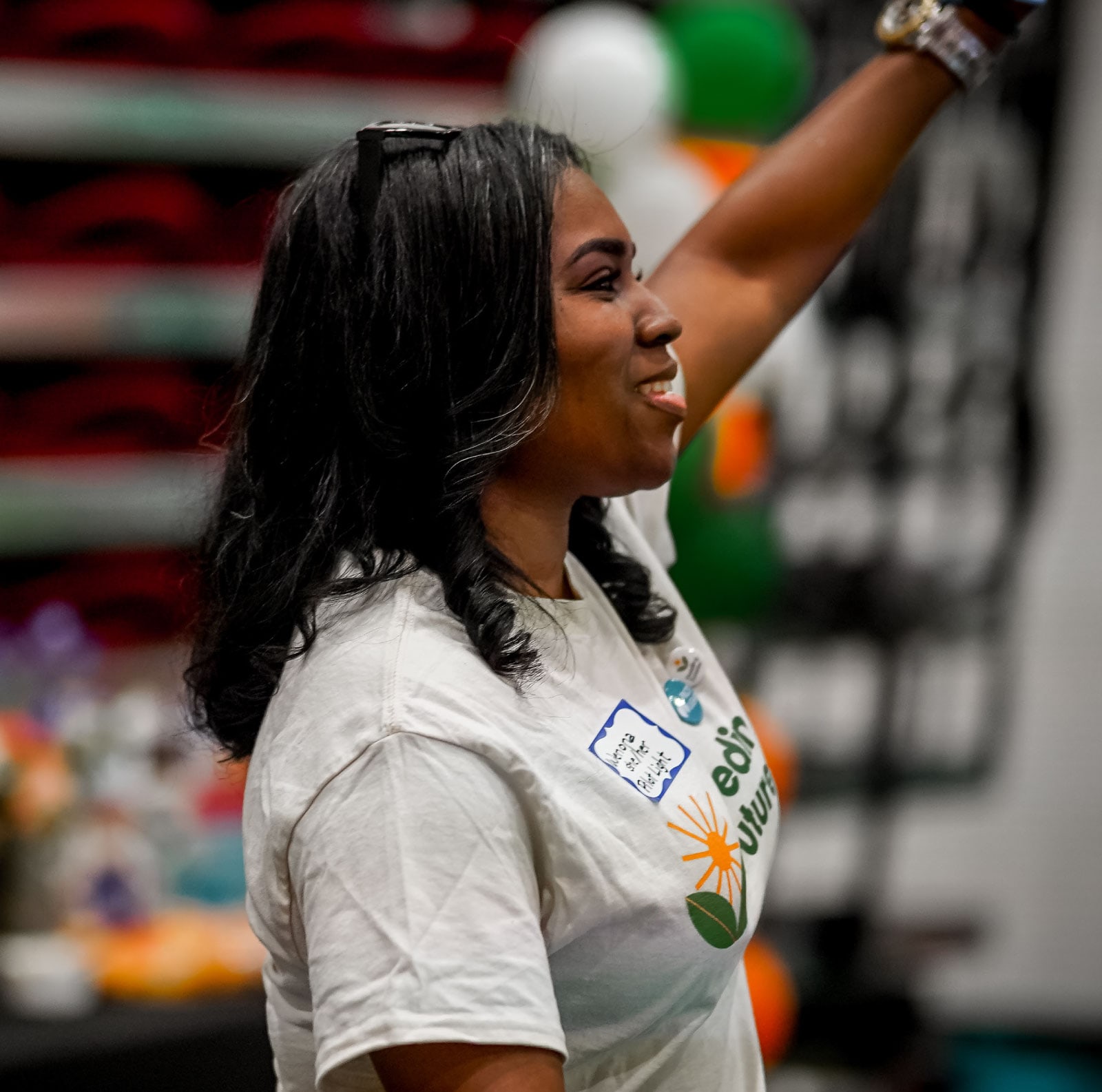 Smiling woman in a white event T-shirt raises her arm overhead at a lively indoor venue, projecting joy, pride, and celebratory energy.