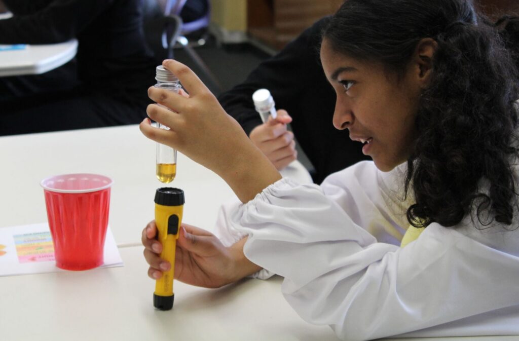 Focused young student in a white lab coat examines liquid in a test tube over a flashlight during a hands-on science experiment.