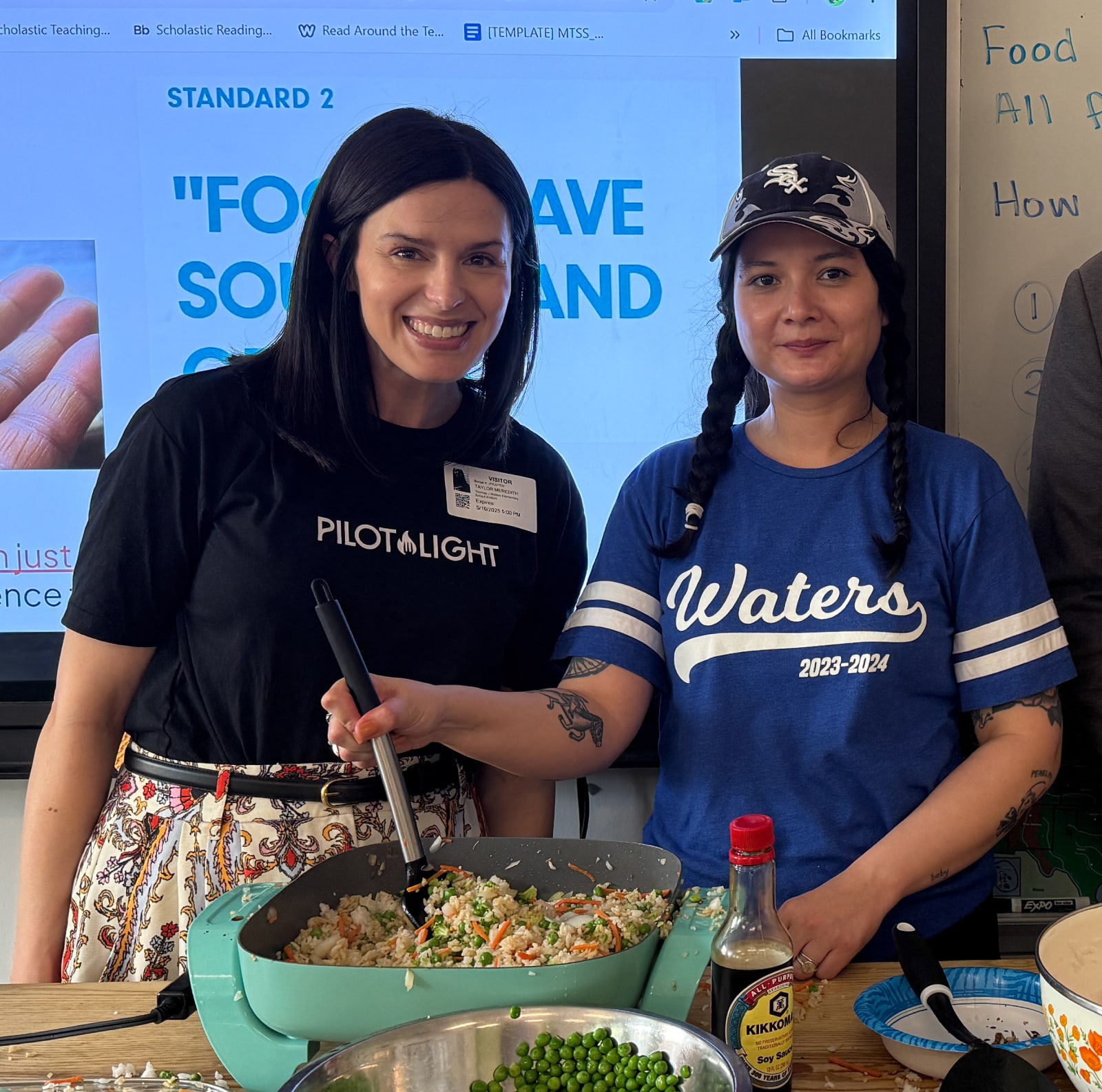Two smiling women pose during a cooking demonstration, holding a pan of rice and vegetables; a presentation screen and appears behind them.